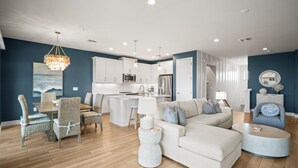 Ein modernes Wohnzimmer mit einem großen weißen Sofa, einem Essbereich mit Korbstühlen und einer Küche mit weißen Schränken und Edelstahlgeräten.