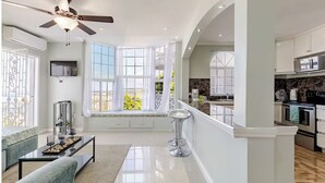 A modern living room with a large window, a ceiling fan, and a kitchen area visible through an open door. - Cozy 1-bedroom apartment in Ocho Rios stunning views of the Caribbean sea. (Ocho Rios)