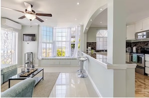 Un salon moderne doté d’une grande fenêtre, d’un ventilateur de plafond et d’un coin cuisine visible à travers une porte ouverte.