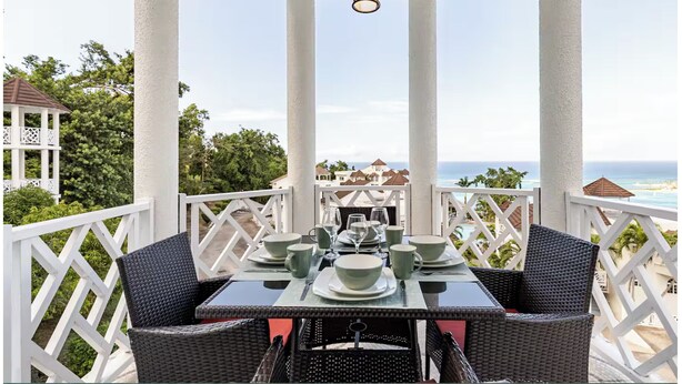 A covered outdoor dining area with a table set for two, white columns, and a view of the ocean. - Cozy 1-bedroom apartment in Ocho Rios stunning views of the Caribbean sea. (Ocho Rios)