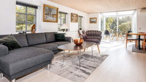 A modern living room with a grey sectional sofa, a round coffee table, and a patterned rug. There are large windows with artwork on the walls, and a dog is visible on the sofa. - 4 Star Holiday Home in Jerup (Jerup)