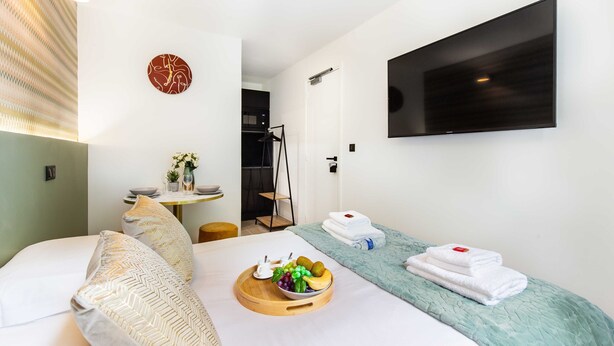 Una habitación de hotel moderna con una cama, una mesita de noche con un frutero, un televisor de pantalla plana en la pared y una pequeña zona de estar con una silla y una mesa.