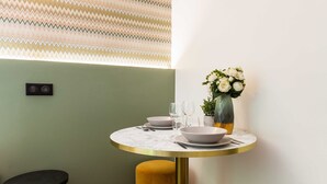 Un área de comedor con una mesa redonda, un otoman amarillo, un reloj de pared y un jarrón con flores.