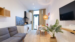 A modern living room with a grey sofa, a dining area with a table and chairs, a kitchen with a stove and washing machine, a TV mounted on the wall, and a potted plant. - Travel Habitat Ruzafa Loft 1 (Valencia / València)
