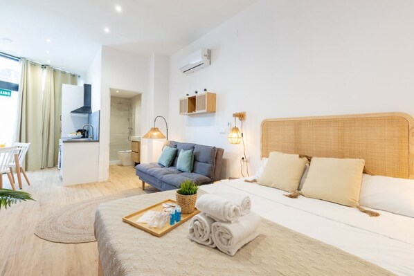 A modern hotel room with a bed, a sofa, a small kitchen area, and a bathroom.