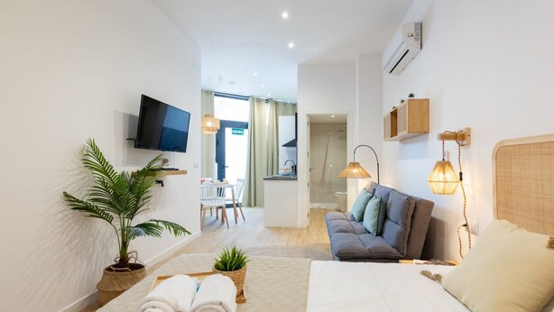 Una habitación de hotel moderna con una cama grande, un sofá, un televisor y un área de comedor visible a través de una puerta abierta.