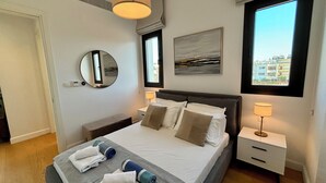 A neatly made bed with pillows, a bedside table with a lamp, a wall-mounted mirror, and a window with a view of buildings.