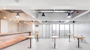 Een moderne, ruime kamer met een houten bank en tafels, grote ramen en hangende pendelverlichting.
