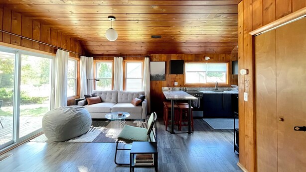 A cozy cabin interior with a wooden ceiling, a sofa, a dining table, and a kitchen area. - Modern Cabin Higgins Lake; walk to Hiking, XC Ski, River, Town, near golf + ORV (Roscommon)