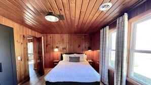 A wooden-paneled bedroom with a bed, two bedside lamps, a window with curtains, and a door leading to another room. - Modern Cabin Higgins Lake; walk to Hiking, XC Ski, River, Town, near golf + ORV (Roscommon)