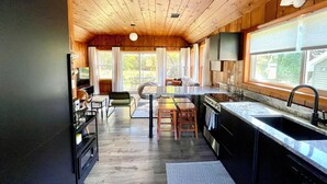A modern kitchen with wooden ceiling beams, a large window, and a dining area with a table and chairs. - Modern Cabin Higgins Lake; walk to Hiking, XC Ski, River, Town, near golf + ORV (Roscommon)