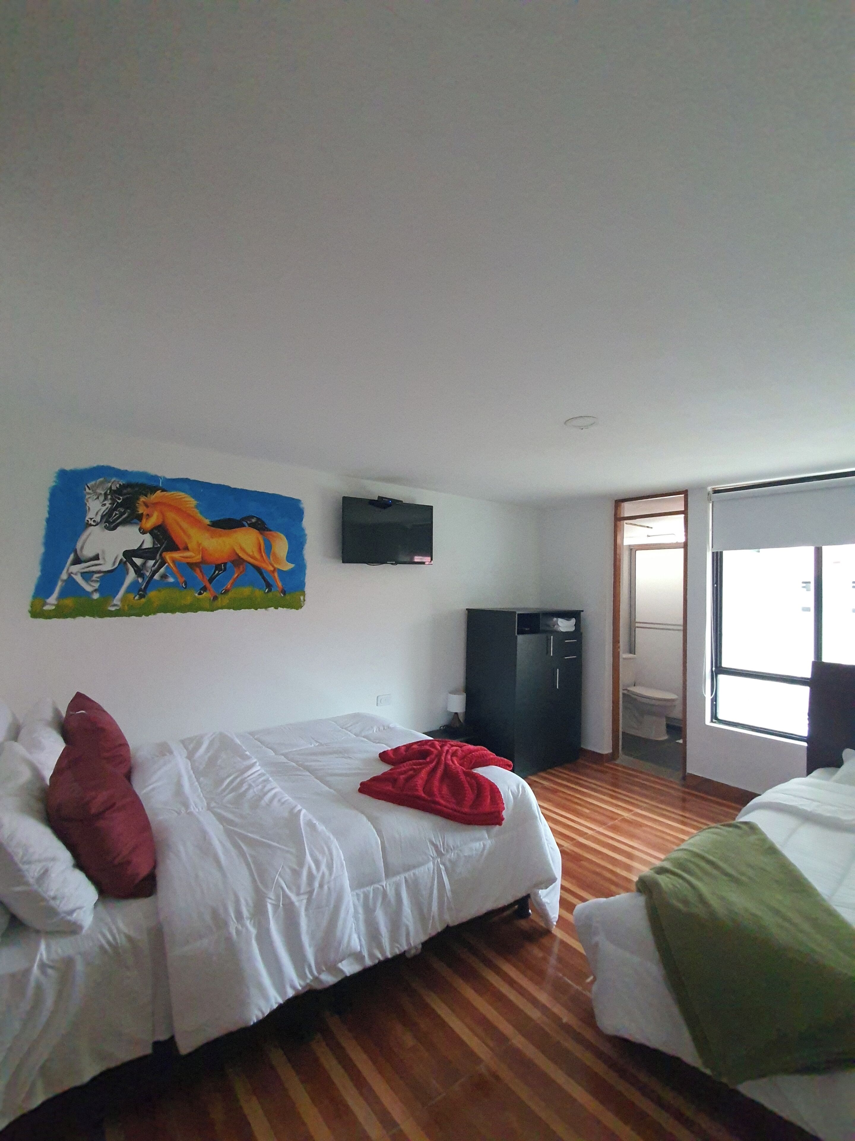 A hotel room with two beds, a TV on the wall, a painting of horses, and a wooden floor.