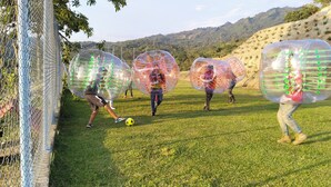 Un campo cubierto de césped con personas dentro de esferas transparentes, una cerca de malla metálica y un paisaje montañoso al fondo.