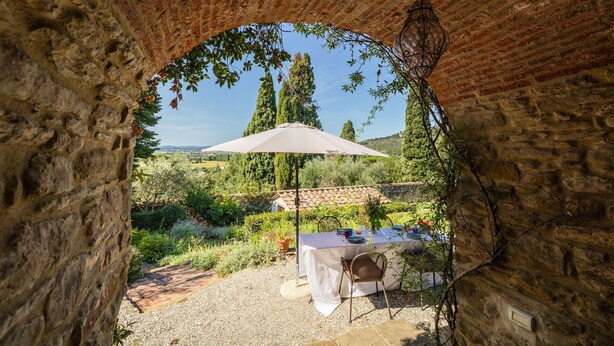 Ein rustikaler Steingewölbepassage führt zu einer Außenbereich-Esszone mit Tisch und Sonnenschirm, umgeben von viel Grün und Blick auf ferne Hügel.