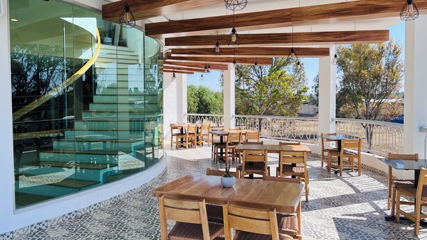 Un espace de restauration extérieur abrité, avec des tables et des chaises en bois, un escalier en verre et une vue sur les arbres et les bâtiments.