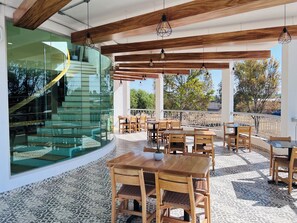A covered outdoor dining area with wooden tables and chairs, a glass staircase, and a view of trees and buildings. - Hotel Mamá Dolores (Dolores Hidalgo)