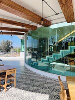 Un interior moderno con vigas de madera, suelo estampado y pared de vidrio que ofrece vistas de unas escaleras exteriores y vegetación.