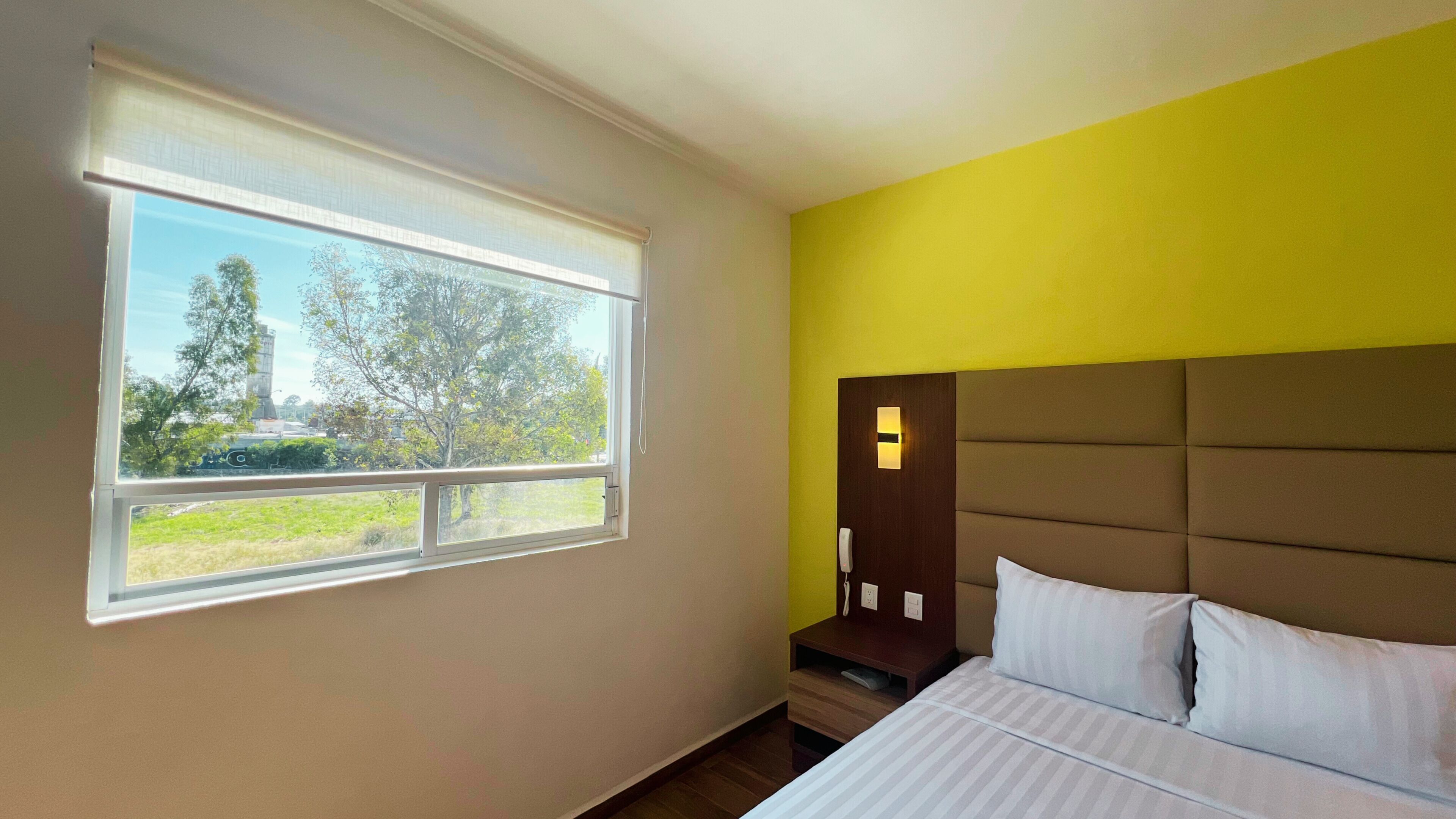A bedroom with a bed, a nightstand, a bedside lamp, and a window with a view of trees and buildings.