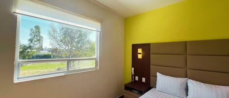 A bedroom with a bed, a nightstand, a bedside lamp, and a window with a view of trees and buildings.