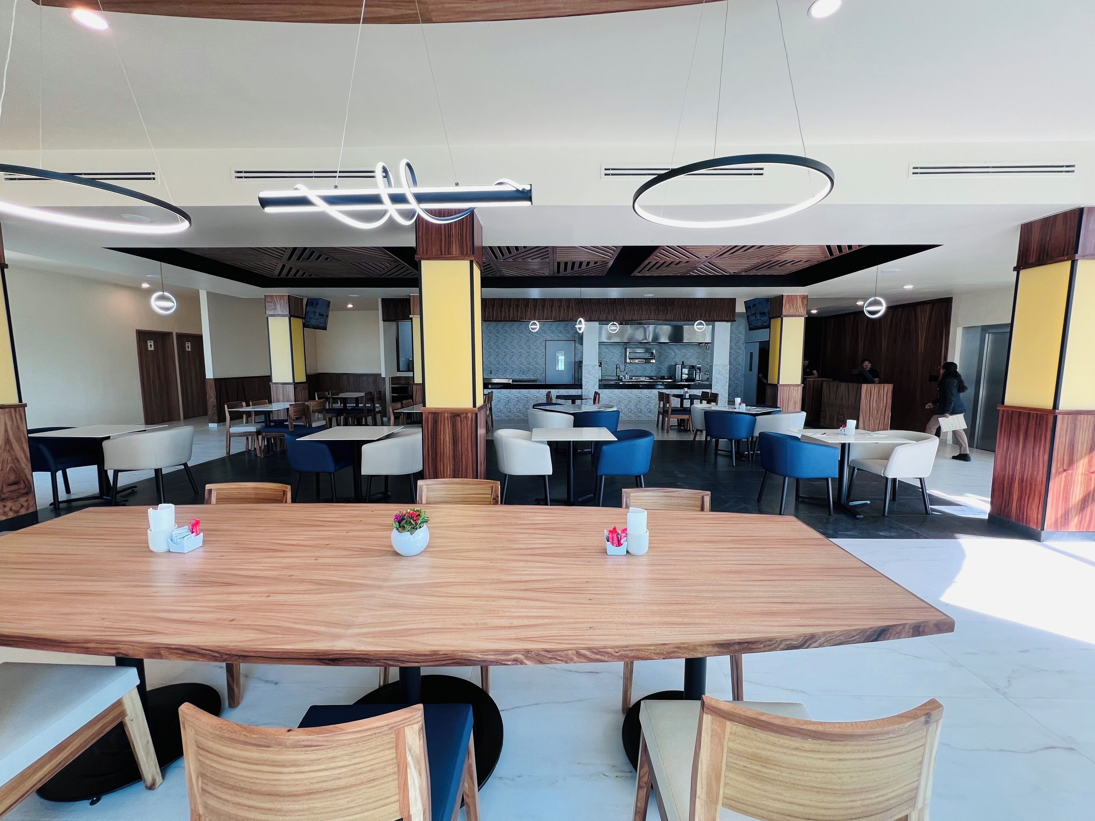 A modern dining area with wooden tables and blue chairs, a kitchen in the background, and a reception desk.
