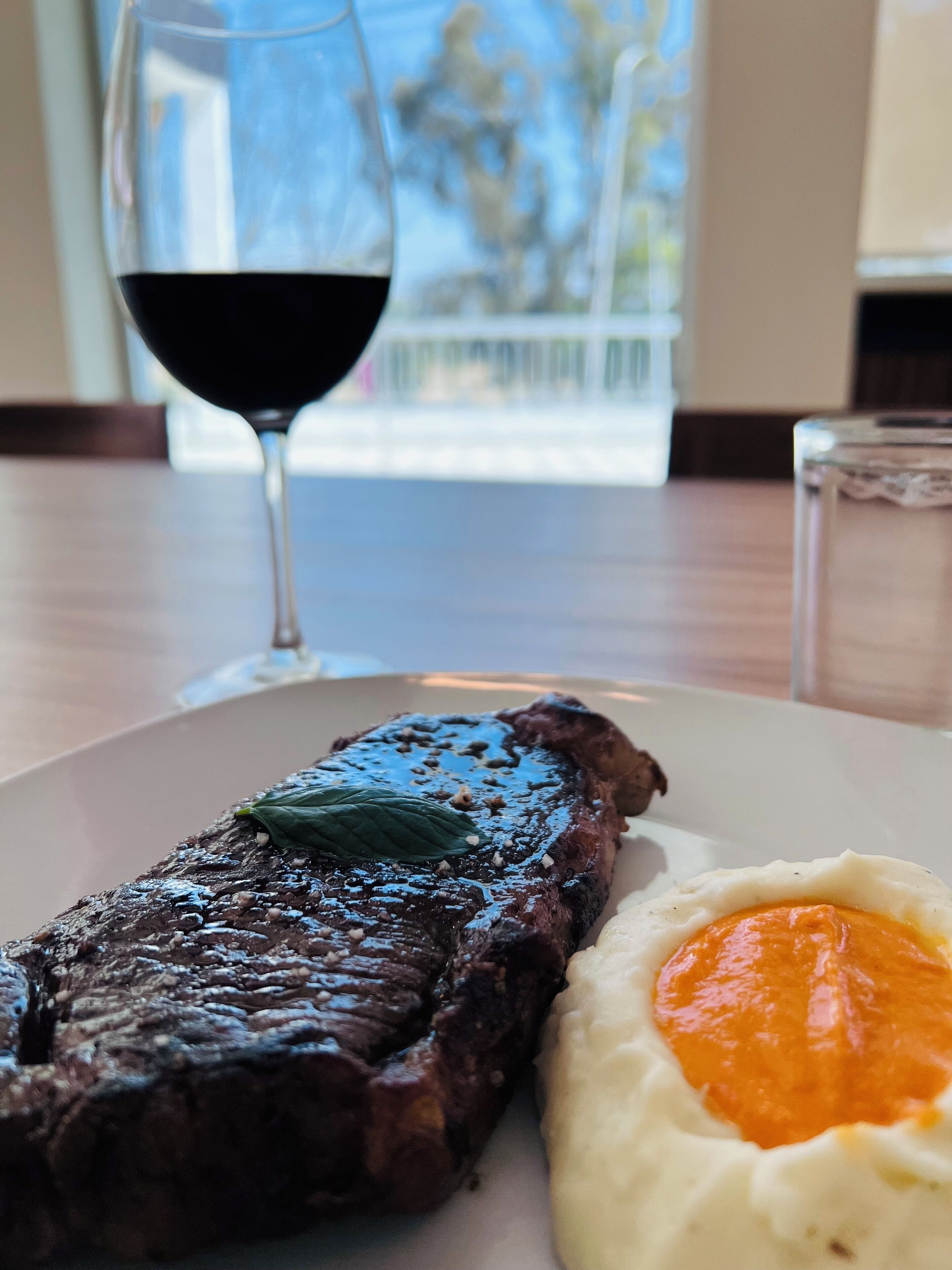 A steak with a garnish and mashed potatoes on a plate, with a glass of red wine and a window view in the background.