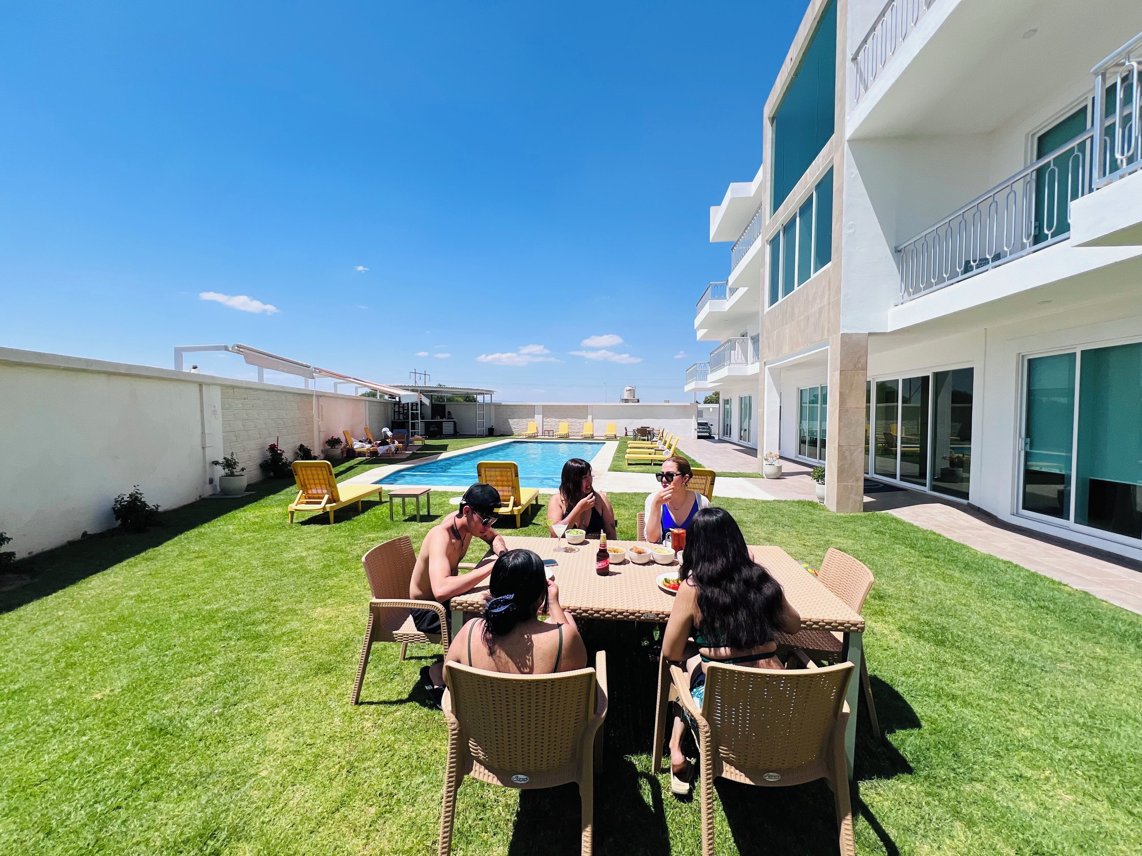 Eine Gruppe von Menschen speist an einem Tisch im Freien, im Hintergrund ist ein Pool zu sehen.