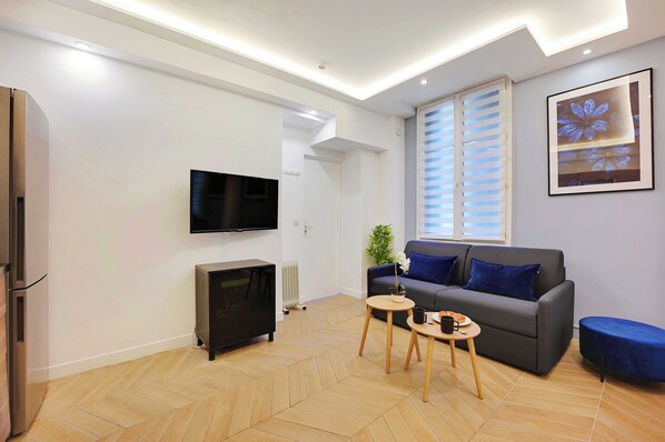 A modern living room with a grey sofa, a flat-screen TV, a small wooden table, and a blue ottoman. - Serene Apartment - 1br/4p - Place de la Bastille (Paris)