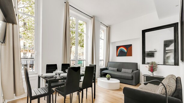 Una sala de estar moderna con comedor, ventanas amplias, un sofá negro, una mesa de centro blanca y un sillón negro.