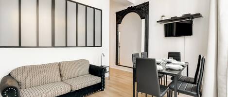 A modern living room with a black and white patterned sofa, a dining table set for four, and a mounted flat-screen TV.