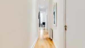 Un pasillo estrecho con suelo de madera y paredes blancas, que conduce a una habitación con una puerta y un radiador.