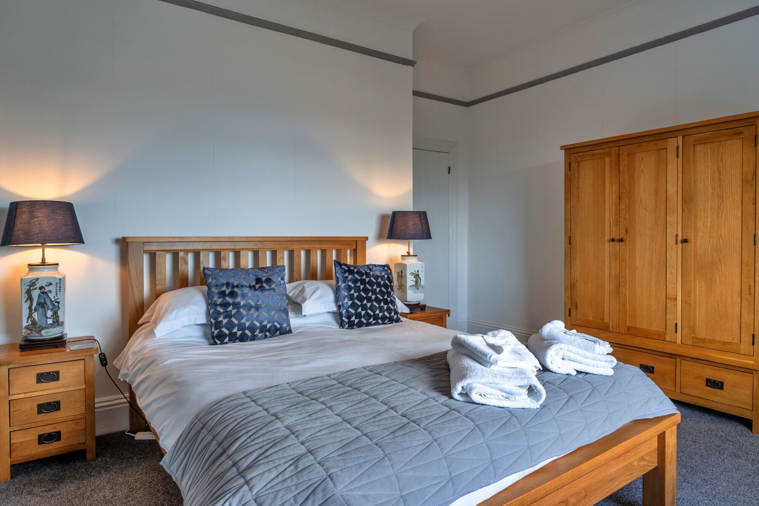 A neatly made bed with a quilted grey bedspread, two bedside lamps, and a wooden wardrobe.