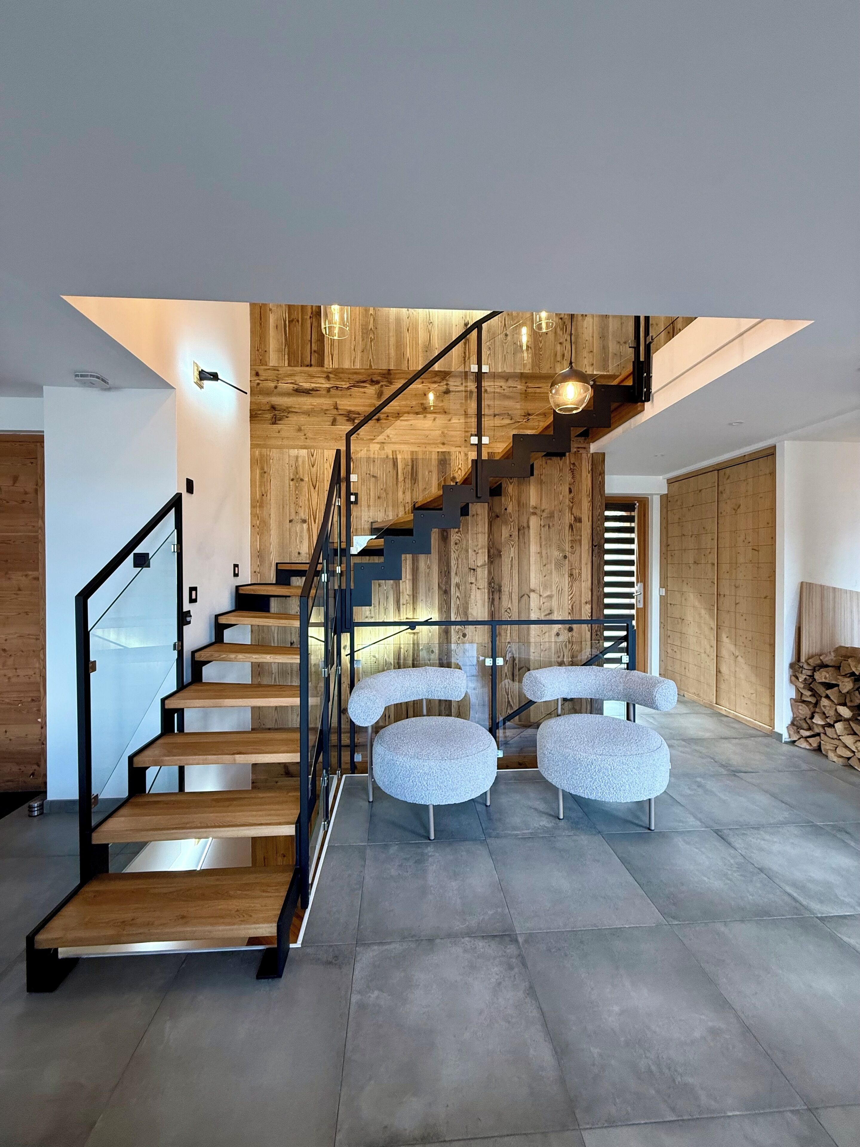 Una escalera moderna con escalones de madera y barandillas de metal, que conduce a un altillo con techo de madera y lámparas colgantes.