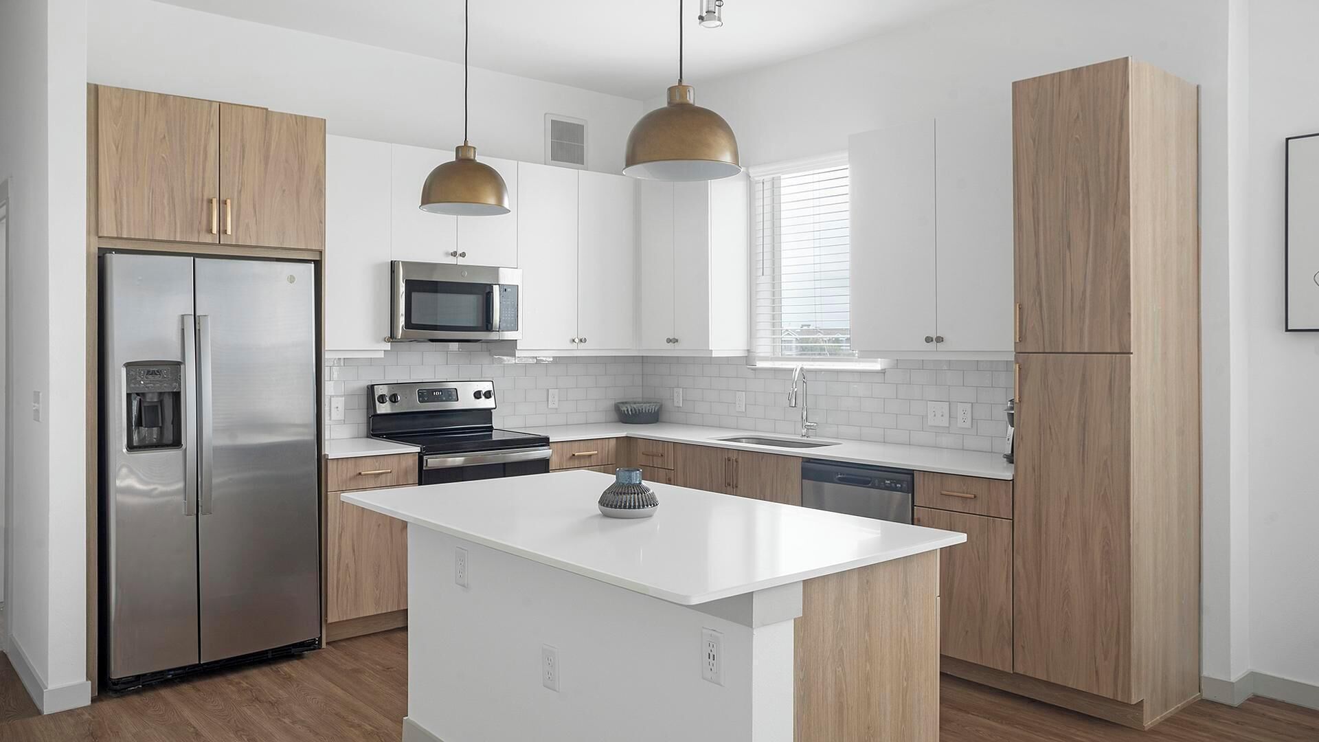 Eine moderne Küche mit Edelstahl-Kühlschrank, Mikrowelle, Herd und Geschirrspüler. Holzschränke und eine weiße Arbeitsplatte vervollständigen das Bild.