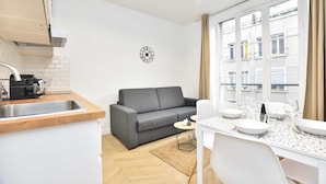 A modern kitchen with a grey sofa, dining table set for four, and a large window with a view of buildings. - Calm Apartment -1br/4p- Bastille (Paris)