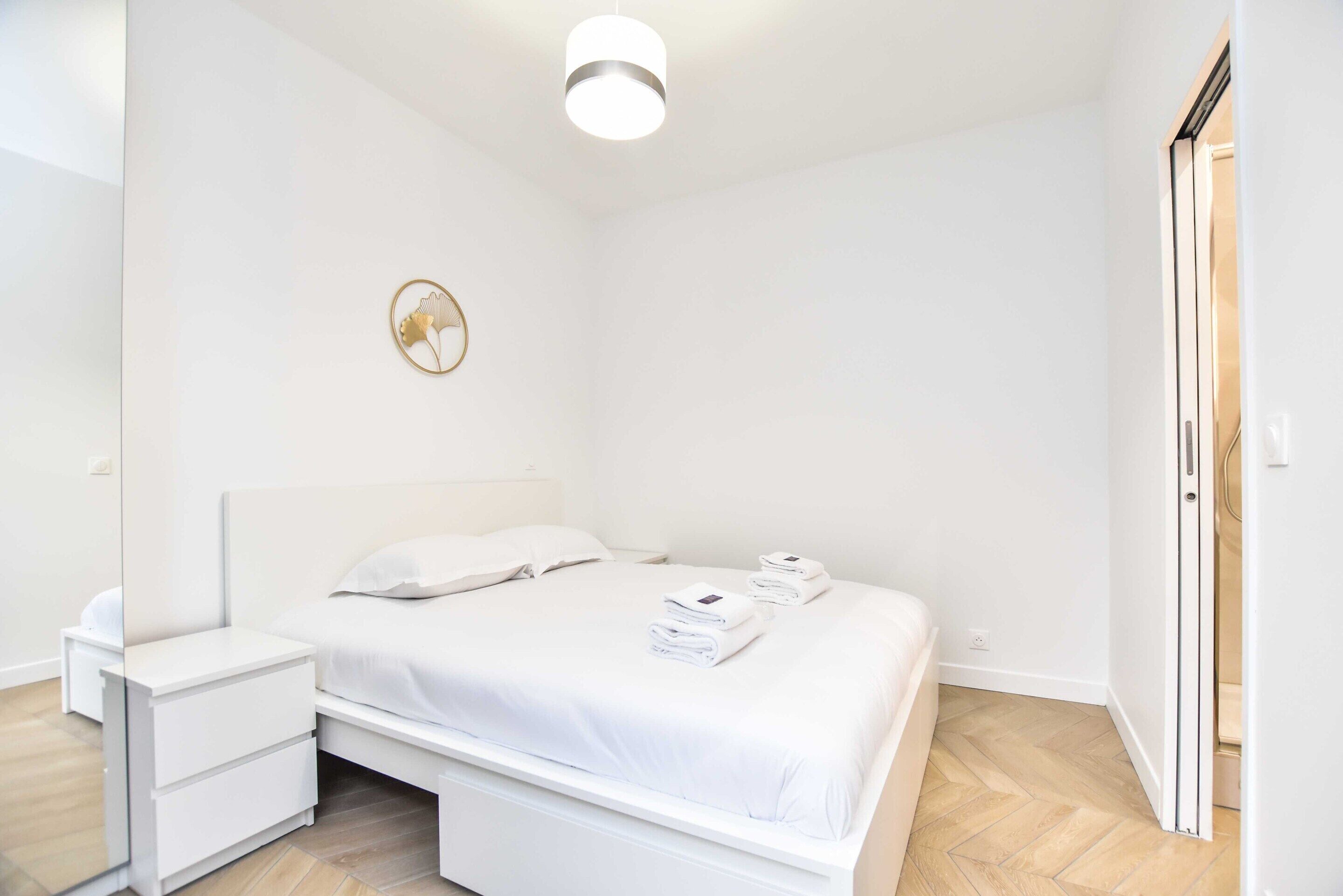 A clean, white bedroom with a bed, bedside tables, and a wall-mounted light fixture.