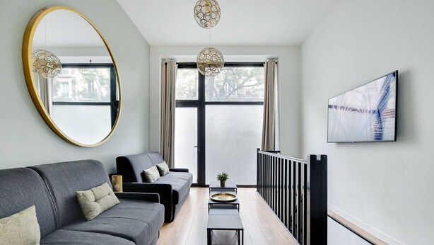 Un moderno salón con un sofá gris, un espejo redondo, un televisor de pantalla plana y suelo de madera.