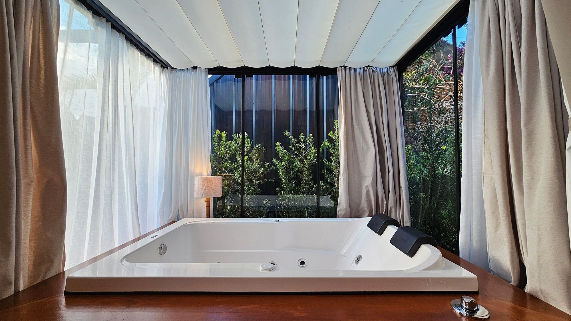 A modern bathroom with a large bathtub, a wooden floor, and a view of greenery through large windows.