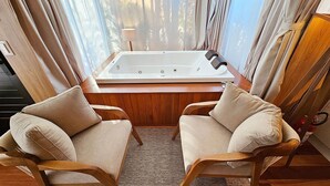 Une pièce avec un plancher en bois, deux fauteuils beiges, une grande baignoire blanche et une fenêtre avec des rideaux.