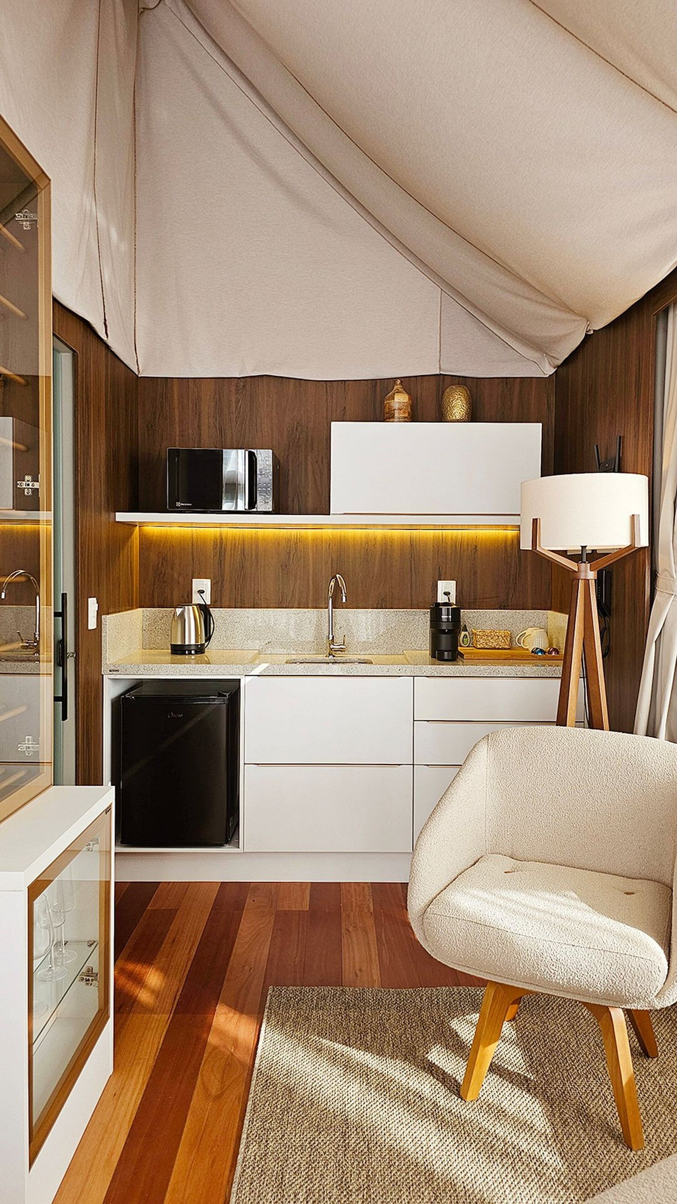 A compact kitchen with a microwave, coffee maker, and a small refrigerator. There's a white chair and a lamp in the room.