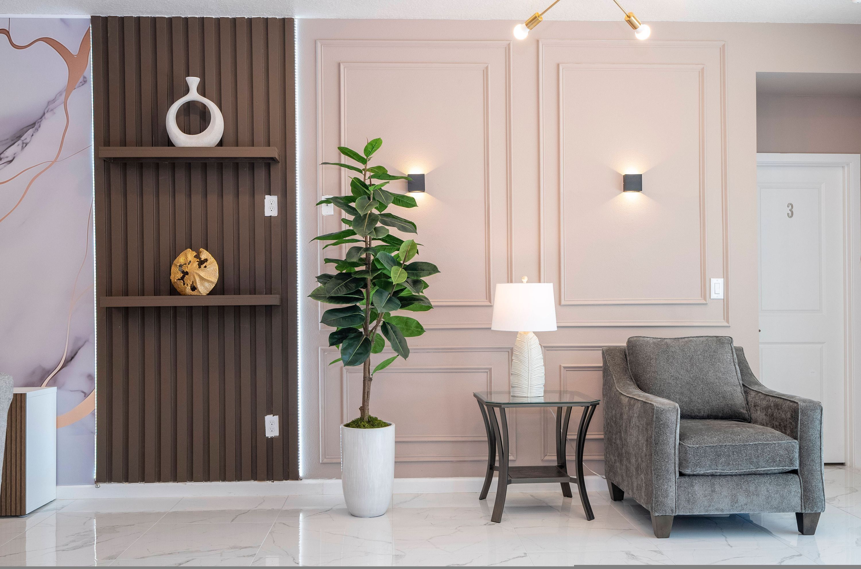 A modern living room with a grey armchair, a potted plant, a small table with a lamp, and a wall-mounted shelf with decorative items.