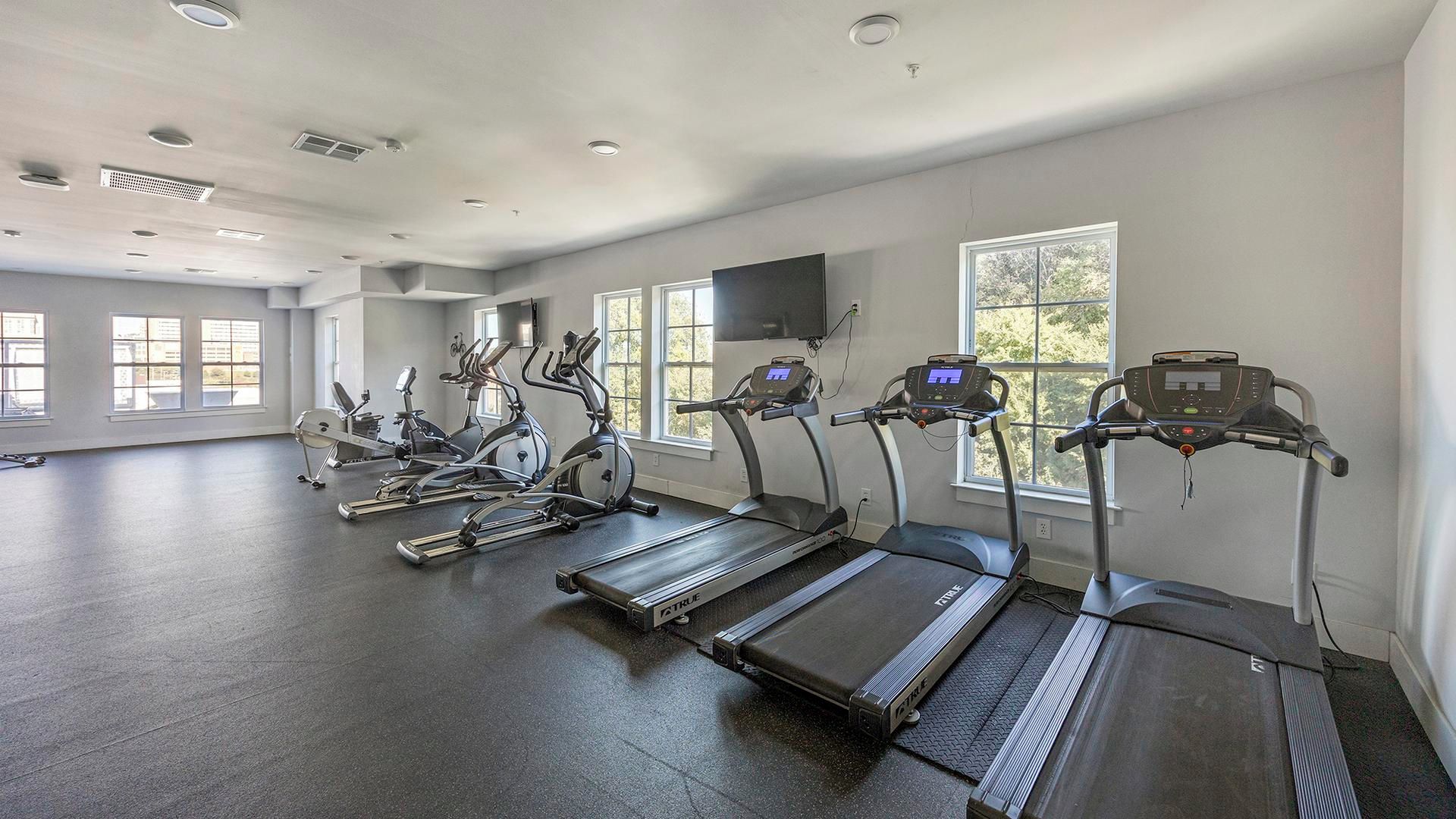 Un gimnasio con cintas de correr, elípticas y bicicletas estáticas, ventanas amplias y suelo plano.