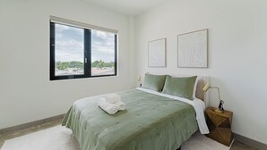 A bedroom with a large window, a bed with green bedding, two framed artworks, and a bedside lamp.