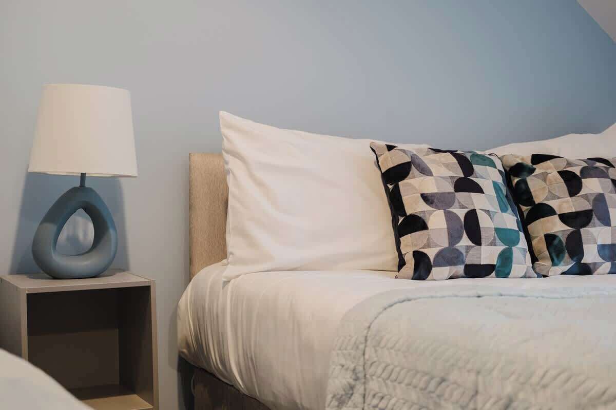A neatly made bed with a white comforter and two patterned pillows, a bedside lamp on a nightstand, and a plain wall.