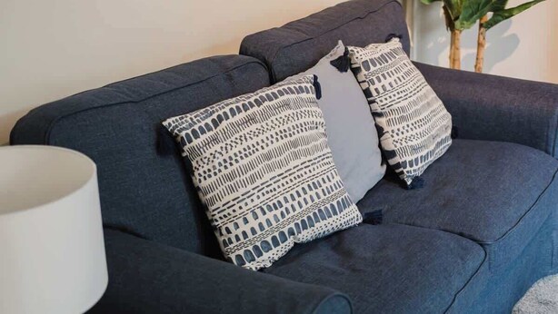 A dark-colored sofa with two patterned cushions and a white lamp. - Spacious 3 Bedroom House Sleeps 6 With WiFi (Bedford)