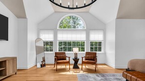 Ein geräumiger Raum mit hohen Decken, großen Fenstern, Holzboden, zwei Ledersesseln, einem Sofa, einem Spiegel und einer Lampe.