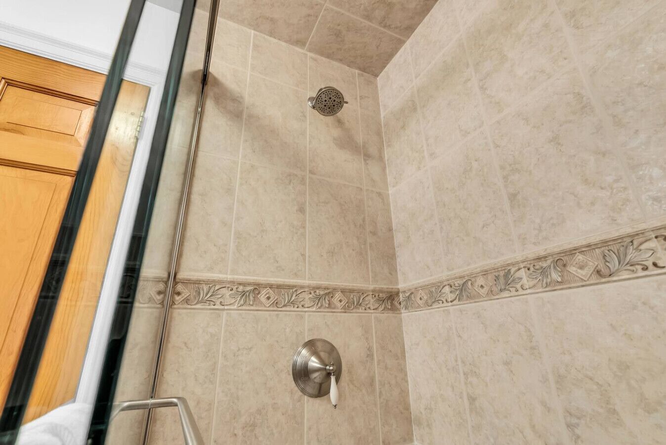 Une douche avec des carreaux de couleur beige, un pommeau de douche et une porte en verre.