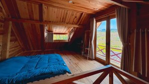 A wooden cabin with a sloped ceiling, a bed with a blue cover, and a view of a red and green landscape through a large window. - Chahill Dalat (Xuan Truong - Da Lat)