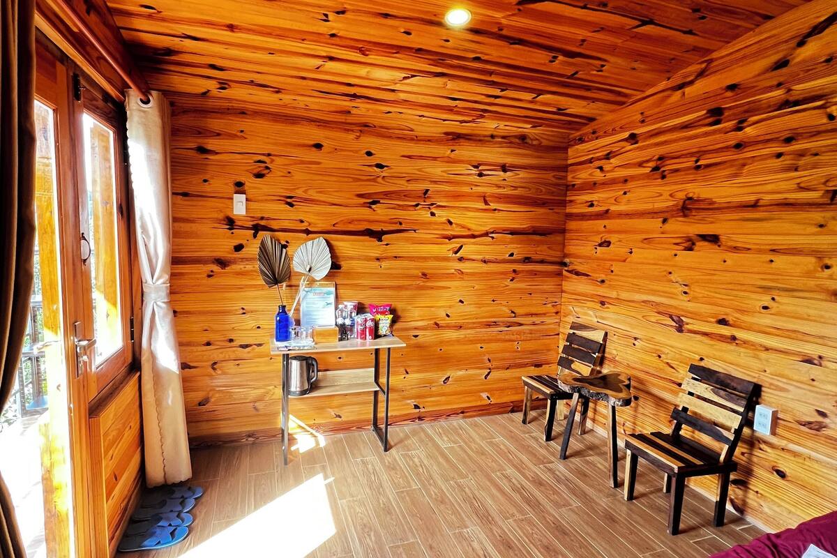 Una cabaña acogedora con paredes y suelos de madera, una cama con manta morada, una mesa con un ventilador y dos sillas.