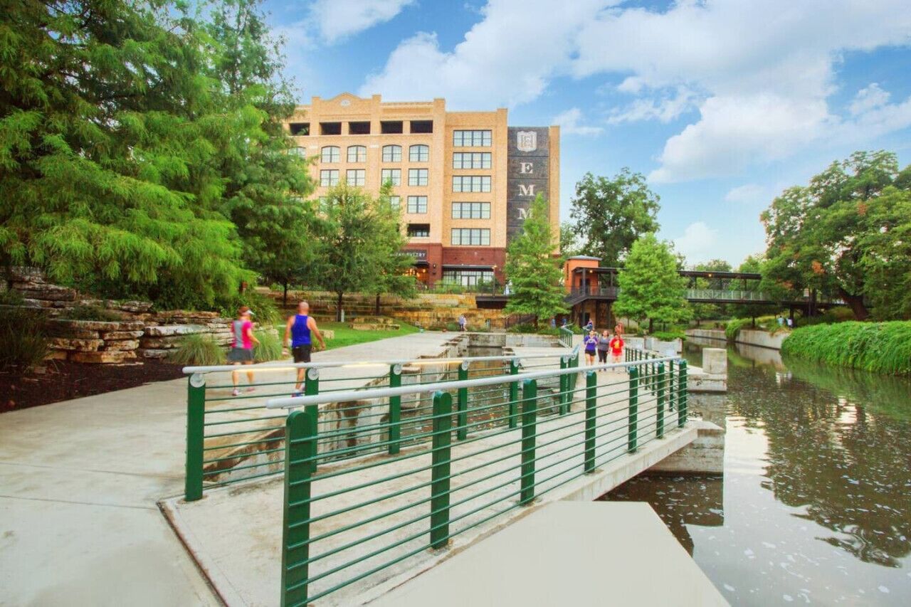 A riverside walkway with a stone wall and greenery, a bridge, and a building with a visible sign.