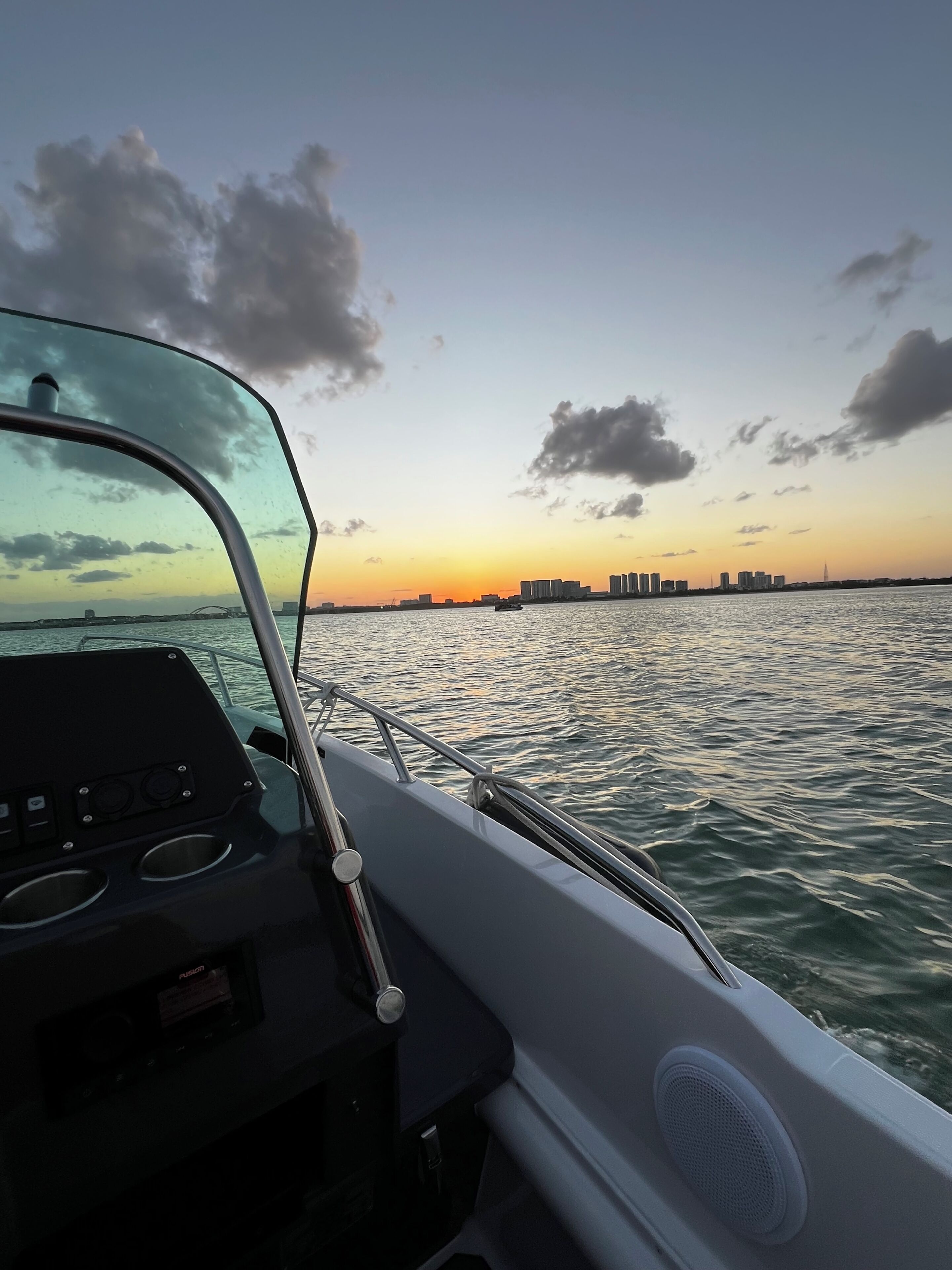 A boat's interior with a control panel and steering wheel, water, and a distant city skyline at sunset.